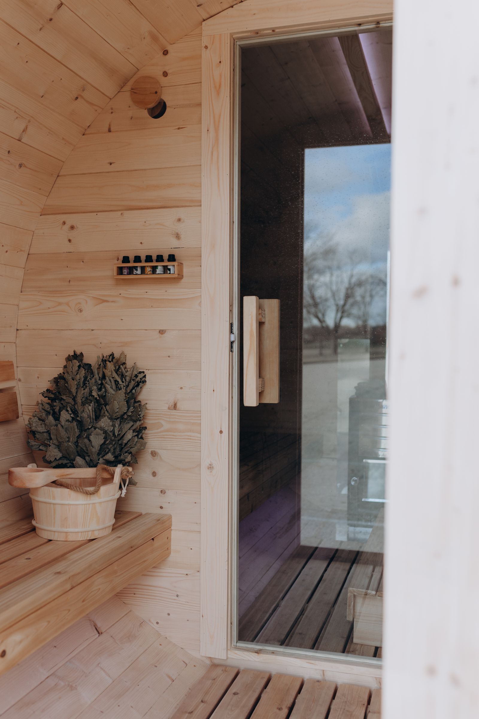 Inside the cedar barrel sauna looking out the glass door, with a whisk and bucket on the bench